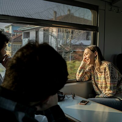 Viaggiatori che godono di un panoramico viaggio in treno, promuovendo la campagna MEININGER Hotels Travel by Train.