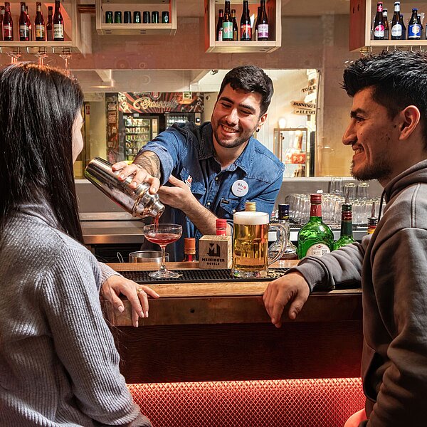 Guests enjoying drinks at MEININGER Hotel Bruxelles City Center bar with a friendly bartender and lively vibe.